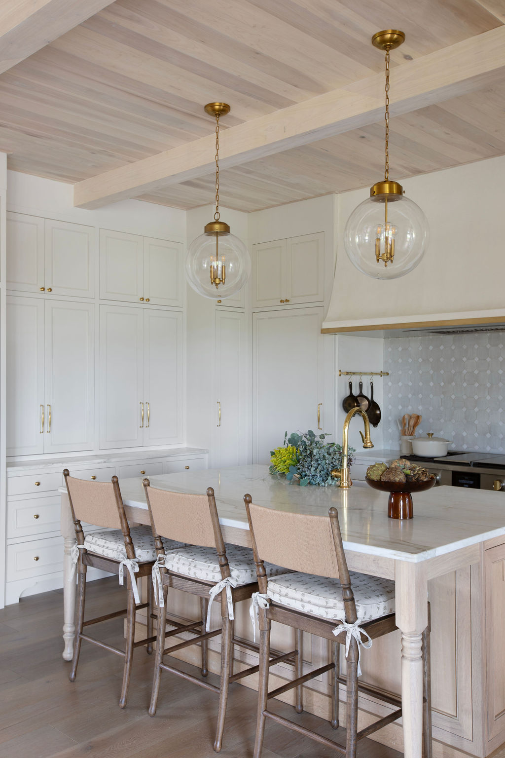 A bright kitchen with pale wood ceiling beams, brass globe pendants, and counter stools with Daffodil in Copper cushions