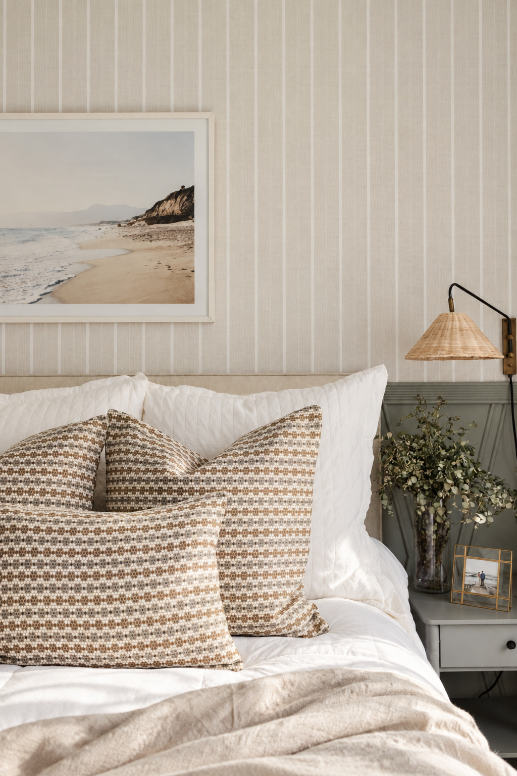 Nectar in Raisin pillows on a bed against striped wallpaper with a woven wall lamp