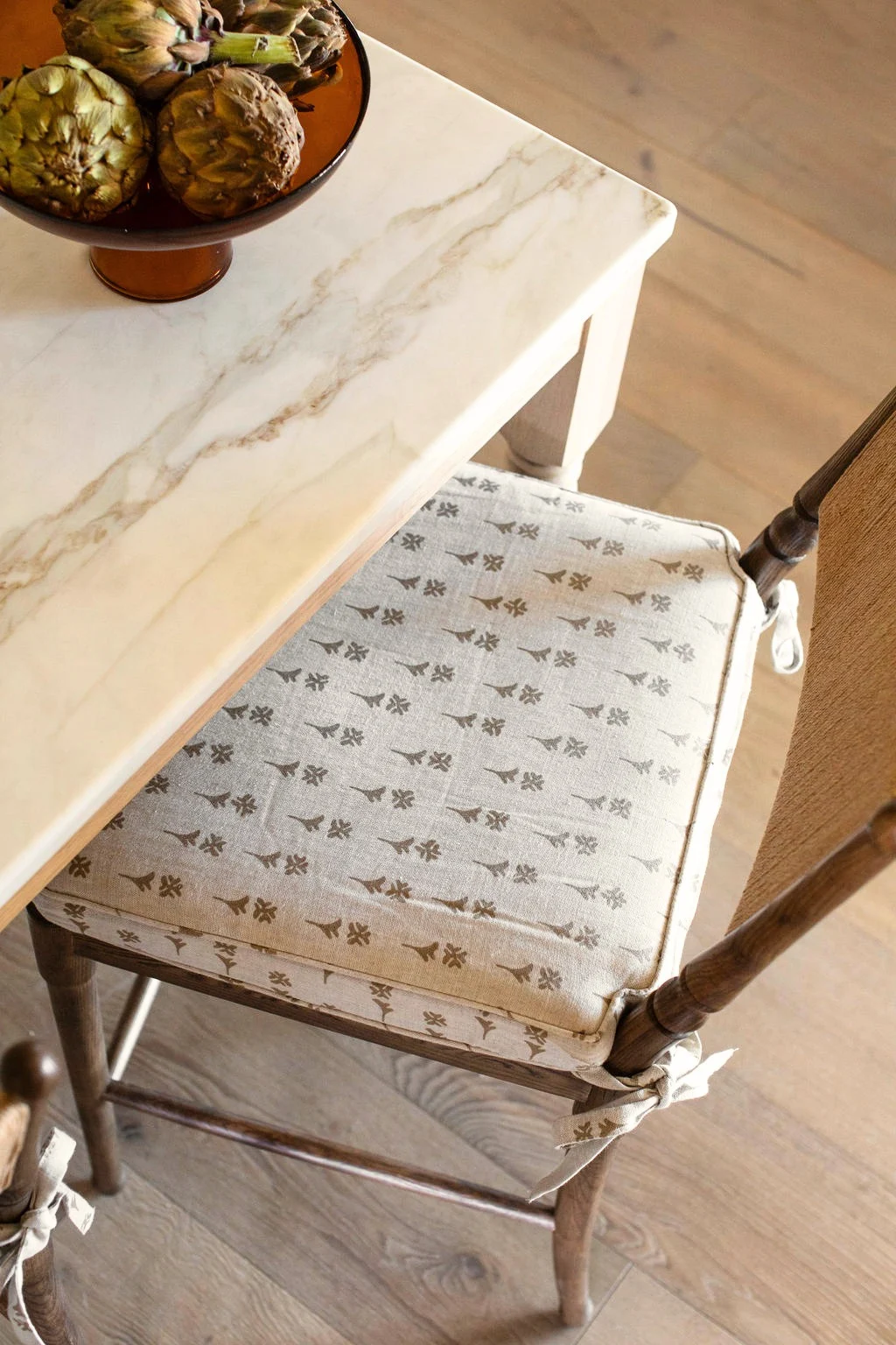A chair cushion in Daffodil in Copper — a small geometric floral in warm brown on natural linen — beside a marble table