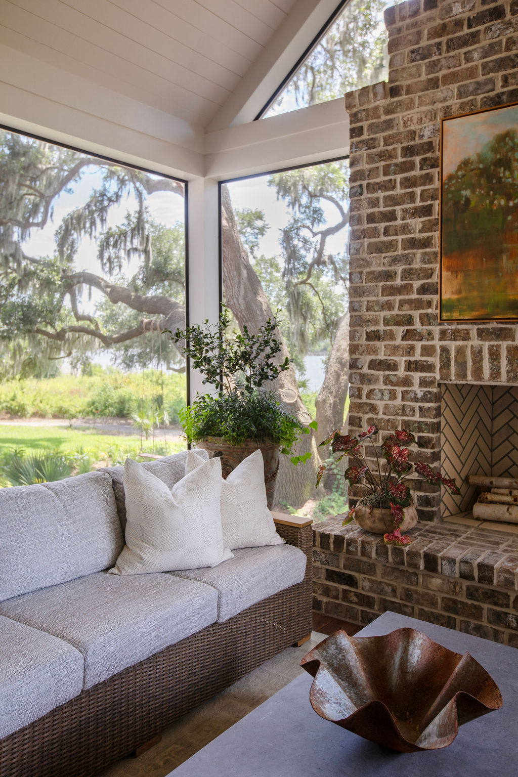 A screened porch with woven seating, neutral cushions, exposed brick and views of live oaks in early autumn light